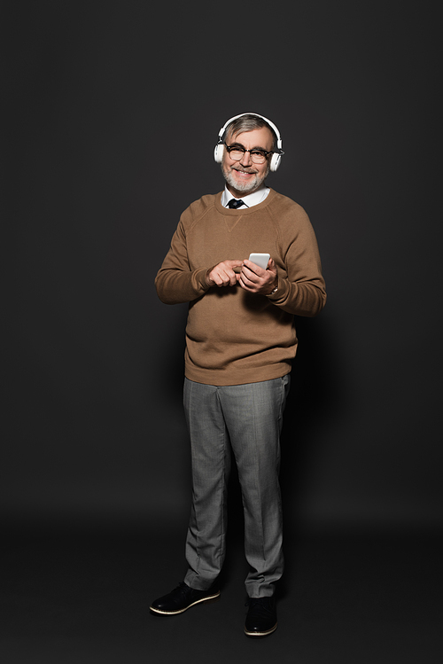full length view of stylish senior man in headphones holding smartphone and smiling at camera on dark grey
