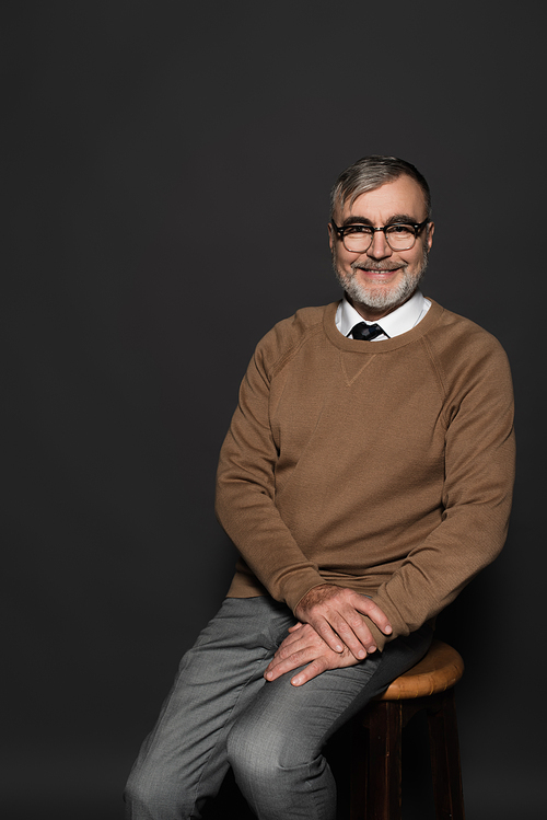 senior man in stylish clothes and eyeglasses sitting on stool and smiling at camera on dark grey