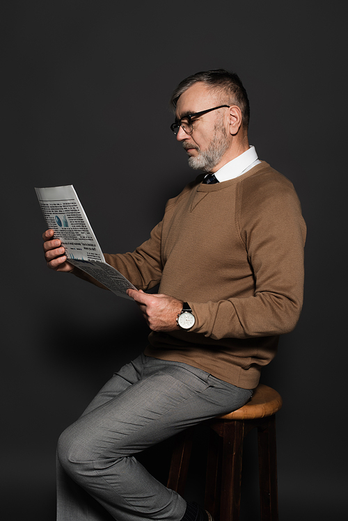 stylish senior man in eyeglasses reading newspaper while sitting on stool on dark grey