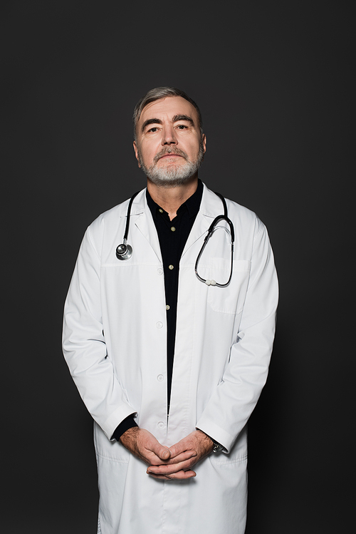 bearded doctor in white coat standing with clasped hands and  isolated on dark grey