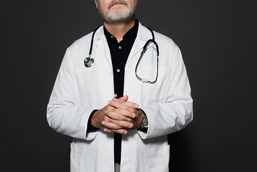 cropped view of senior doctor with stethoscope on neck standing with clasped hands isolated on dark grey