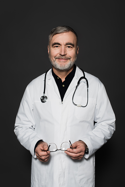 positive bearded physician holding eyeglasses and smiling at camera isolated on black