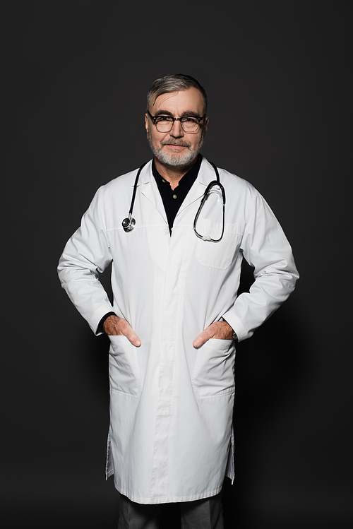 smiling senior doctor  while standing with hands in pockets on dark grey