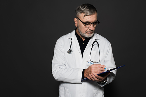 senior physician in eyeglasses and white coat writing on clipboard isolated on black