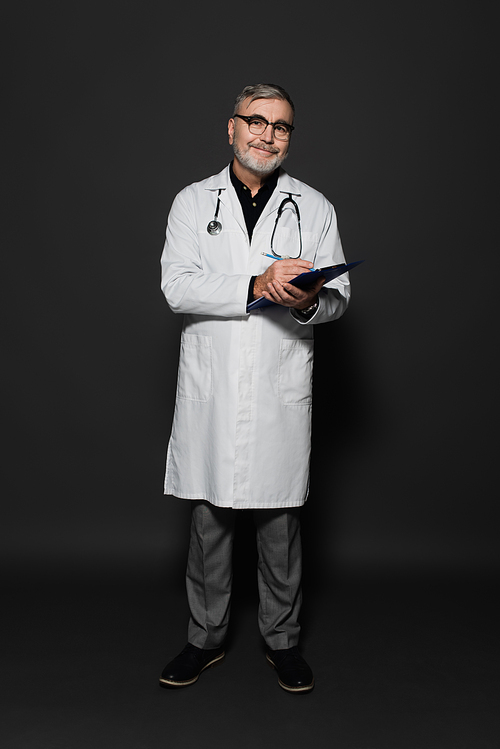 full length view of happy senior doctor in white coat writing on clipboard on dark grey