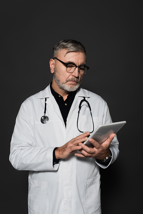 senior doctor in white coat and eyeglasses using digital tablet isolated on black