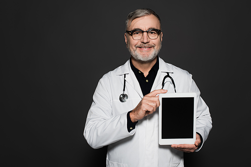 joyful senior doctor in white coat and eyeglasses holding digital tablet with blank screen isolated on black