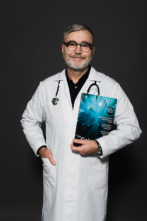 smiling physician in eyeglasses standing with magazine and hand in pocket of white coat isolated on dark grey