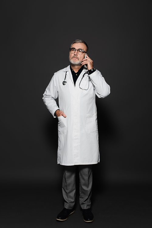 full length view of senior doctor standing with hand in pocket and talking on smartphone on dark grey