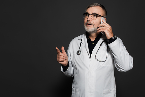 bearded doctor looking away and pointing with finger while talking on cellphone on dark grey