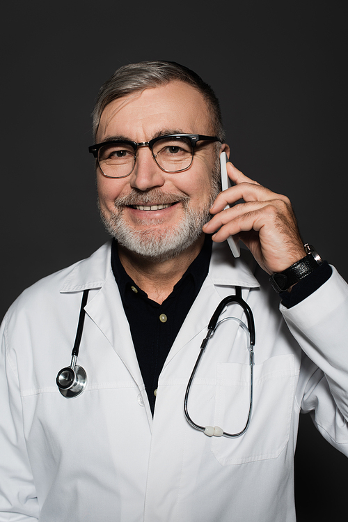 smiling senior doctor in eyeglasses talking on mobile phone isolated on dark grey