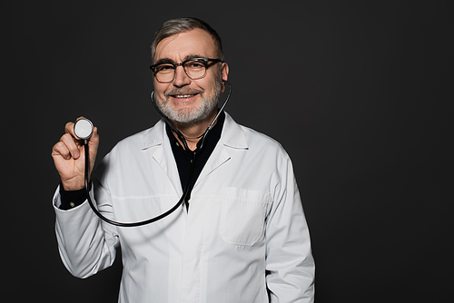 cheerful senior doctor in eyeglasses holding stethoscope isolated on black