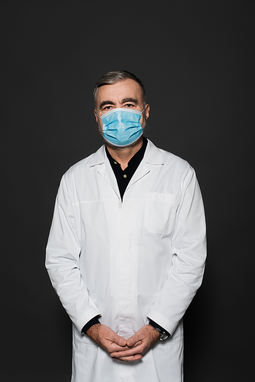 senior physician in white coat and medical mask  isolated on dark grey
