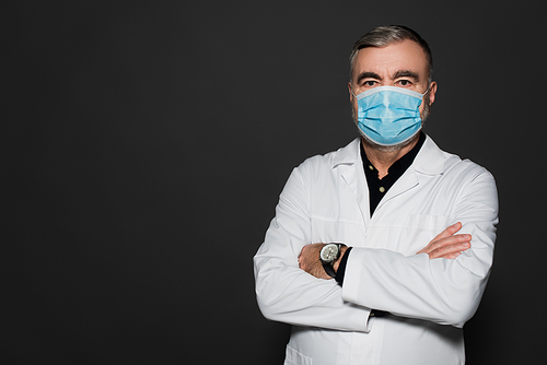 doctor in white coat and medical mask standing with crossed arms and  isolated on dark grey