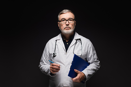 senior doctor in eyeglasses holding pencil and clipboard while  isolated on black