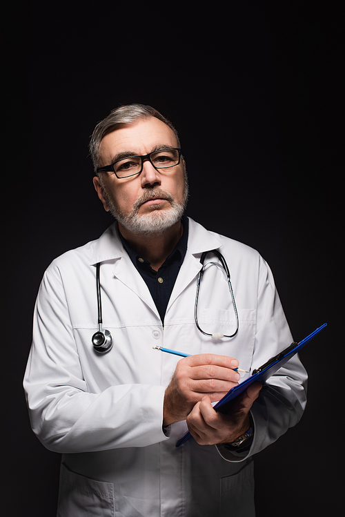 senior doctor in eyeglasses writing on clipboard while  isolated on black