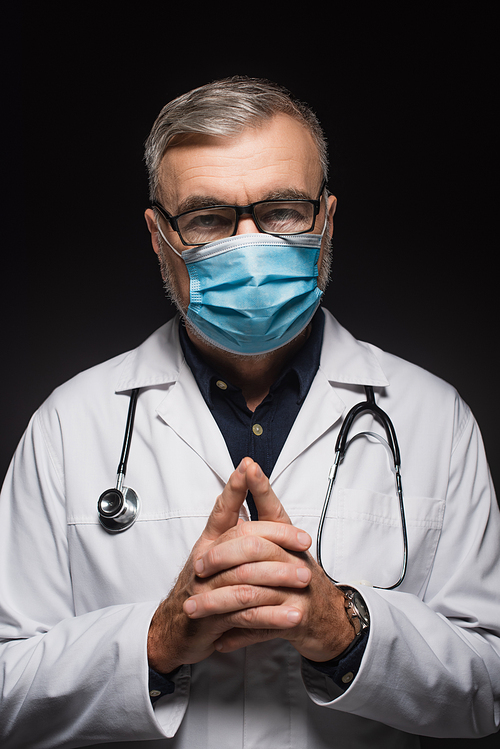 doctor in medical mask and eyeglasses standing with clasped hands and  isolated on black