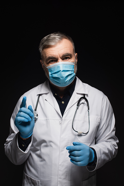 senior doctor in medical mask and latex gloves pointing with finger while  isolated on black
