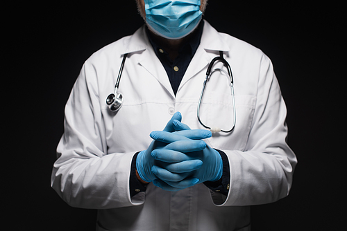 cropped view of doctor in medical mask and latex gloves standing with clasped hands isolated on black