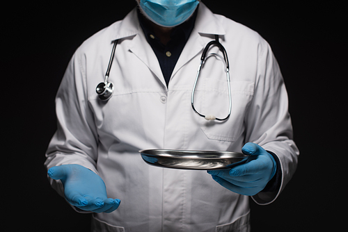 cropped view of doctor in medical mask and latex gloves holding stainless medical tray isolated on black