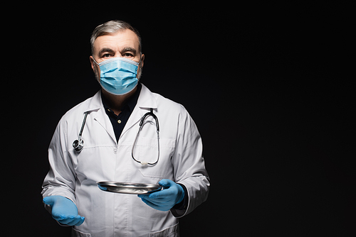senior physician in medical mask holding metallic medical tray isolated on black