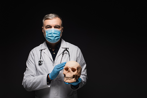 senior physician in medical mask and latex gloves holding skull isolated on black