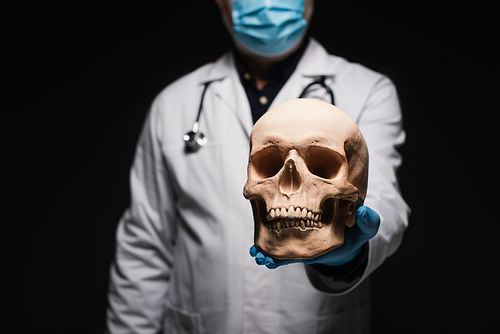 cropped view of blurred doctor in medical mask holding skull isolated on black
