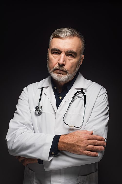 senior bearded doctor with stethoscope on neck standing with crossed arms isolated on black