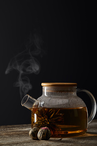 transparent teapot with blooming tea and tea balls on wooden table isolated on black with copy space