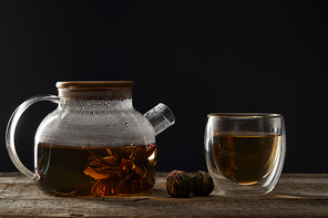 transparent teapot and glass with blooming tea on wooden table isolated on black