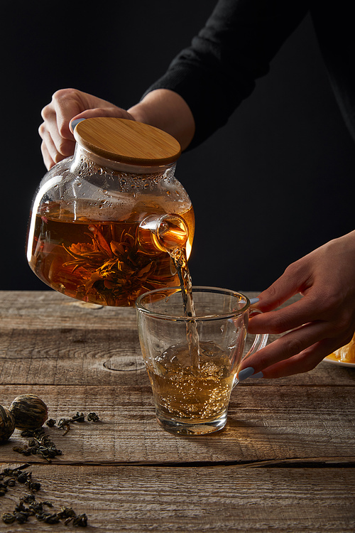cropped view of young woman pouring blooming tea from teapot in cup isolated on black