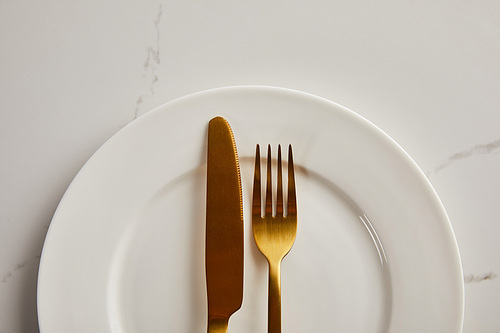 top view of golden knife and fork on clean white plate on marble table