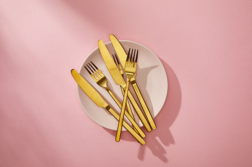 Top view of shiny golden cutlery on empty plate on pink background