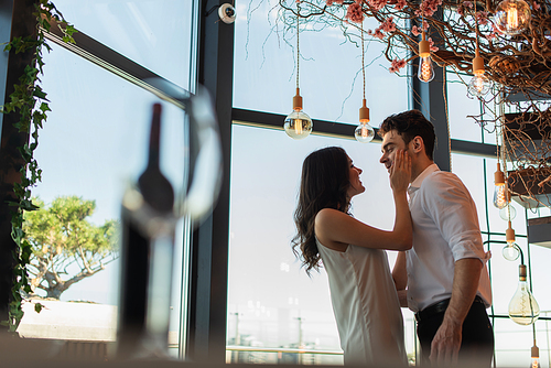 side view of happy woman in slip dress touching face of boyfriend in restaurant