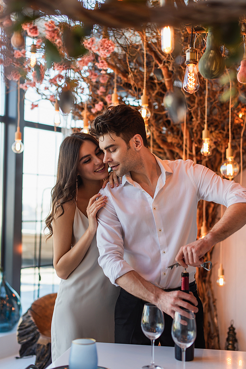 cheerful woman hugging boyfriend opening bottle of red wine in restaurant