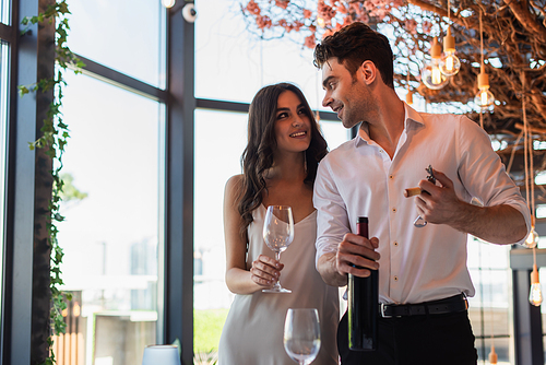 man holding corkscrew with cork near bottle and looking at cheerful girlfriend in slip dress