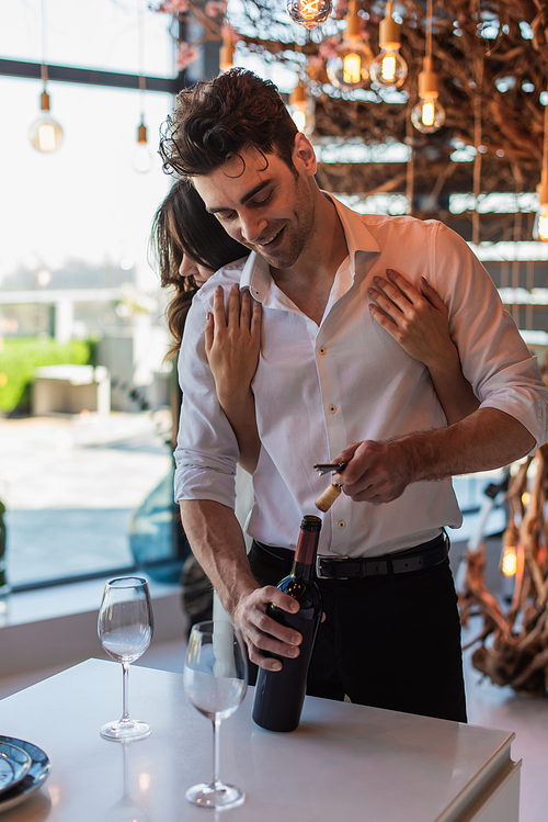 brunette woman hugging happy boyfriend opening bottle of red wine in restaurant