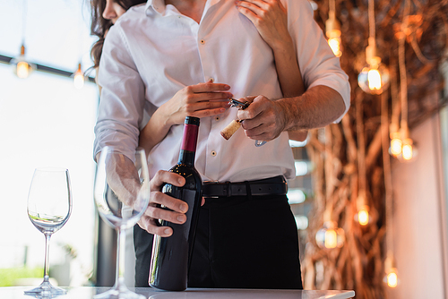 cropped view of brunette woman hugging boyfriend opening bottle of red wine in restaurant