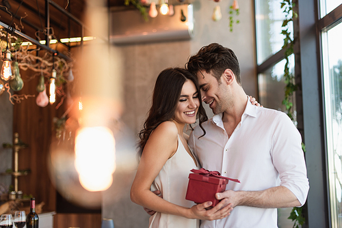 joyful man holding red gift box near smiling girlfriend in slip dress in restaurant