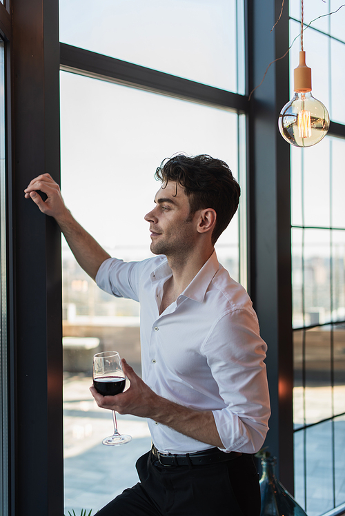 elegant man in white shirt holding glass of red wine while standing near window