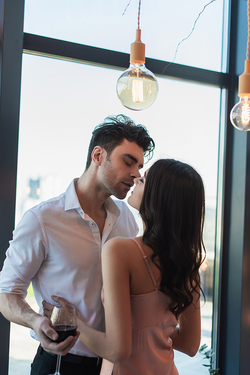 man holding glass of red wine and kissing brunette woman in slip dress