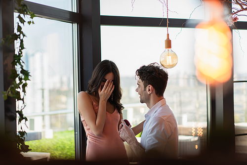 happy man making proposal to girlfriend in slip dress covering face in restaurant