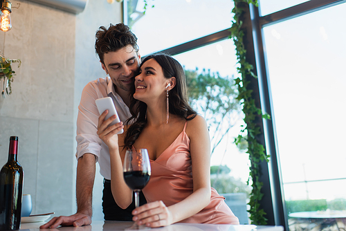 happy couple in wireless earphones looking at each other near smartphone and bottle of wine