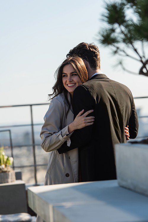 man hugging cheerful woman in coat on terrace
