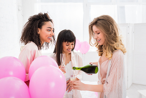 cheerful multicultural girls pouring champagne from bottle into glasses on bachelorette party with pink balloons