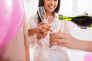 cropped view of happy girls pouring champagne from bottle into glasses on bachelorette party with pink balloons