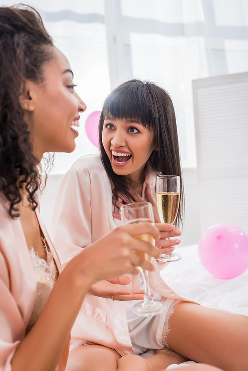 surprised multicultural girlfriends with glasses of champagne talking on pajama party