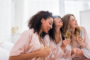 smiling multiethnic girlfriends having fun and holding champagne glasses on pajama party