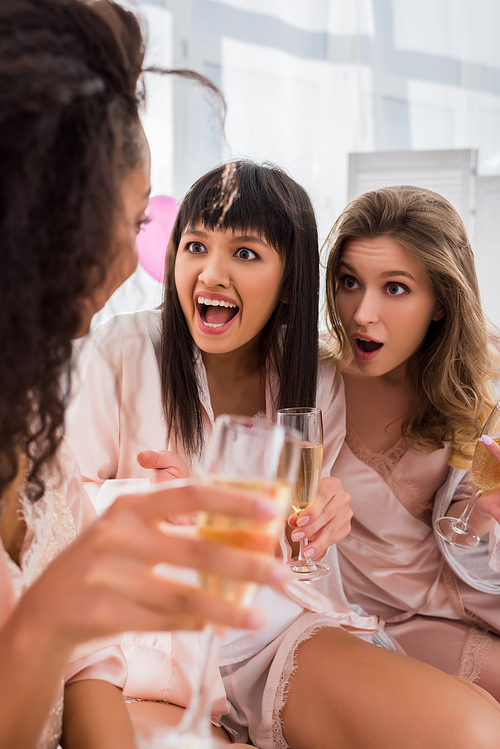 shocked multiethnic girlfriends with glasses of champagne talking on pajama party