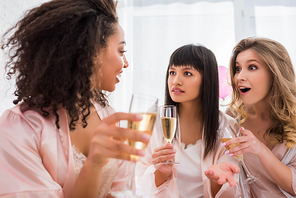 surprised multiethnic girlfriends with glasses of champagne talking on pajama party